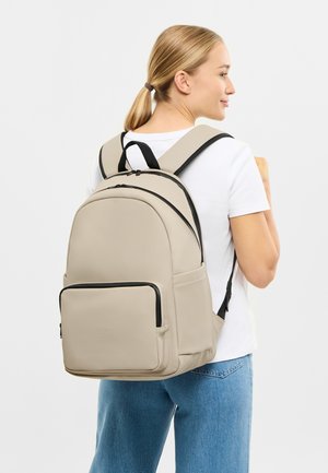 Beige backpack with black accents, smooth material, rounded shape, and a front zip pocket; worn by a person with light hair.