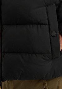 Čierna puffer vesta vyrobená z hladkého, vodoodolného materiálu s prešívaným polstrovaním a textúrovaným povrchom. Obsahuje detail v okrúhlom logu.