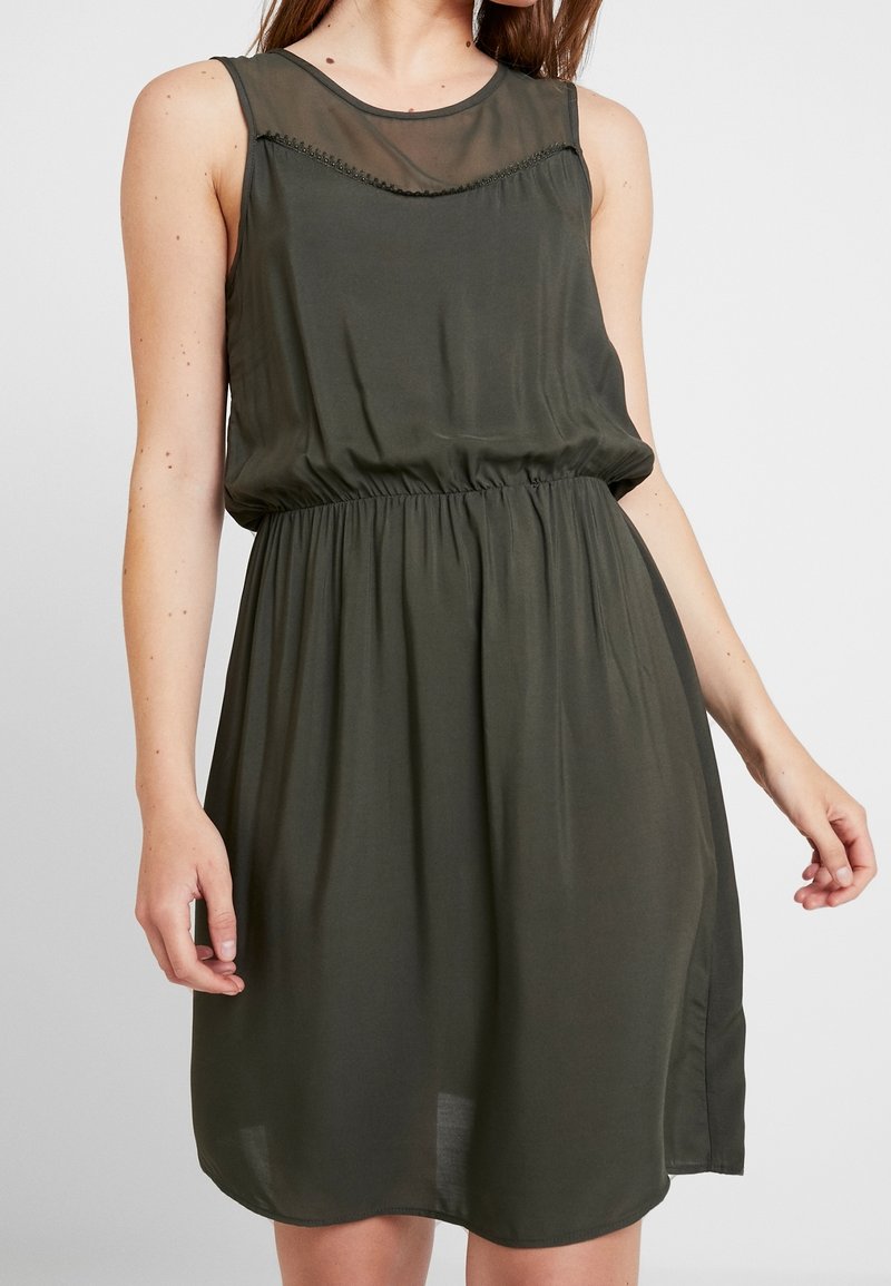 Olivgrön ärmlös klänning med en genomskinlig halsringning, rynkad midja, flödande tyg och en sidoklyvning. Slät textur och avslappnad passform.