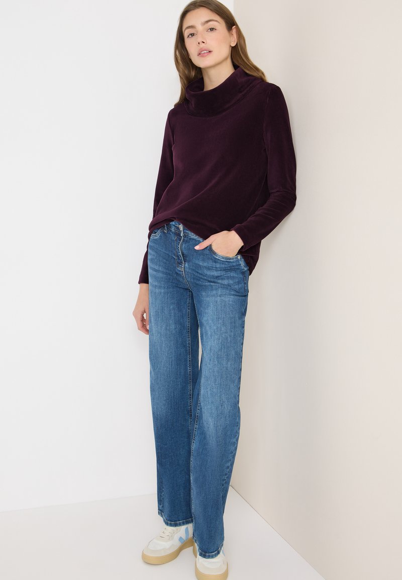 Burgunderfarbener Cowl-Neck-Pullover, kombiniert mit hellblauen weiten Jeans und beigen Sneakers. Glatter Stoff, lockere Passform und klassische Denim-Details.