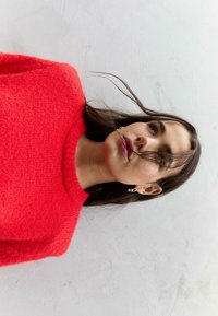 Maglione lavorato a maglia rosso, con scollo rotondo. Orecchini a cerchio argentati. Capelli che coprono parzialmente il viso, su uno sfondo chiaro.