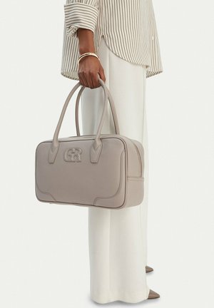 Mujer sosteniendo un bolso de cuero topo con doble asa y logo en relieve, vistiendo una camisa a rayas, pantalones blancos y pulseras doradas.