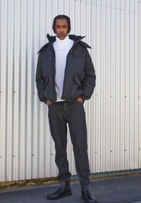 Giacca puffy nera con colletto alto, dolcevita bianco, jeans in denim scuro e stivaletti neri, su uno sfondo in metallo ondulato.