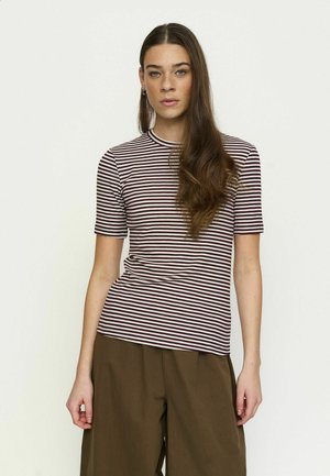 Vrouw met lang bruin haar draagt een aansluitend, kort mouwloos bordeaux-wit gestreept shirt en losse bruine broek, staand tegen een effen achtergrond.
