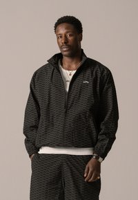 Veste zippée noire avec un motif tonal subtil, col haut et poignets élastiques, portée avec un t-shirt blanc et un pantalon assorti.