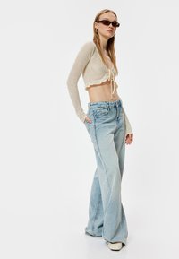Beige gebreide cropped top met lange mouwen en een striksluiting, gecombineerd met baggy lichtblauwe spijkerbroek. Gelaagde materialen en een relaxte pasvorm.