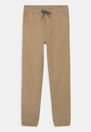 Beige bukser med elastik i taljen og snøre, sidelommer og elastiske manchetter ved anklerne, lagt fladt på en hvid baggrund.