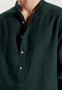 Camicia in lino verde scuro con colletto, placca stile henley e bottoni argentati. Tessuto strutturato visibile.