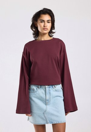 Femme portant un haut ample à manches longues bordeaux avec des manches larges et une mini-jupe en denim bleu clair devant un fond uni.