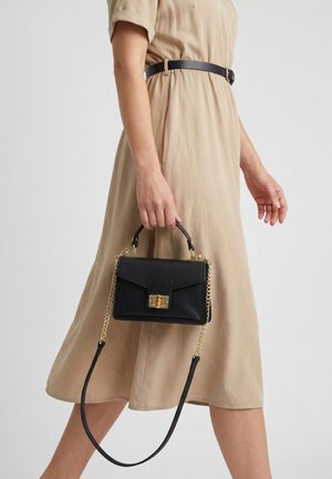 Frau in beigem Kleid mit schwarzem Gürtel hält eine schwarze Lederhandtasche mit goldener Kette und Verschluss, während sie vor einem schlichten Hintergrund geht.