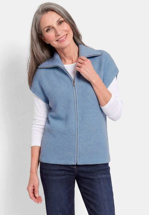 Lächelnde mittelalte Frau mit grauen Haaren, die einen blauen ärmellosen Reißverschluss-Pullover über einem weißen Langarmshirt und dunklen Jeans trägt.