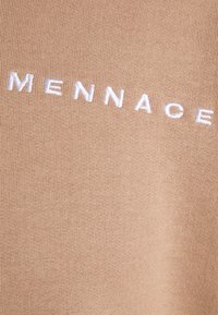 Beige látka s měkkou texturou, na které je bílý vyšívaný text "MENNACE" napříč přední stranou.