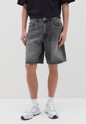 Shorts en denim gris foncé avec une coupe décontractée, ourlet effiloché, poches avant et fermeture à bouton. Associés à des chaussures de sport blanches et des chaussettes.