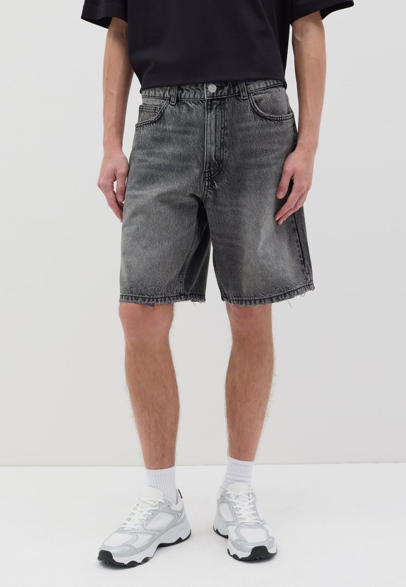 Shorts de mezclilla gris oscuro con un corte relajado, dobladillo deshilachado, bolsillos frontales y cierre de botón. Combinados con zapatillas deportivas blancas y calcetines.