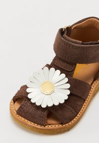 Sandale en velours marron avec un accent floral décoratif. Présente une grande marguerite blanche avec un centre jaune. Design ouvert à l'avant et sangle ajustable.