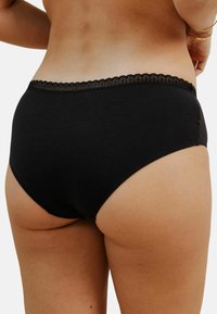 Femme portant des sous-vêtements en coton noir avec des bordures en dentelle, vue de dos de la taille vers le bas sur un fond blanc.