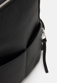 Mustaksi teksturoidusta nahasta valmistettu reppu, jossa on vetoketjullinen yläosa, sivutasku ja tukeva olkahihna. Materiaalissa on pikkukivimäinen pinta.