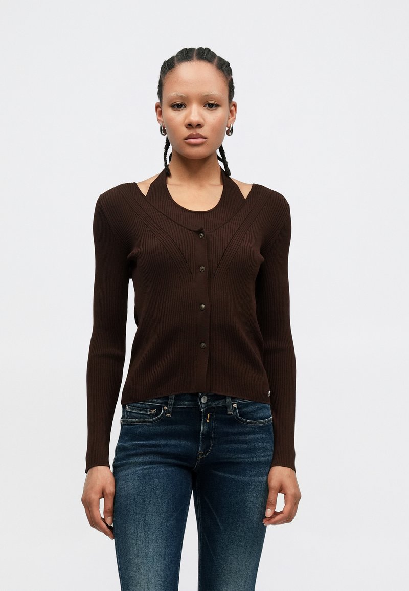 Brown ribbed cardigan with a round neck, button front, and cut-out shoulder design. Paired with dark blue fitted jeans.