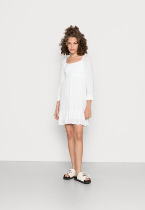 Vestido branco com decote quadrado, mangas bufantes e corpete drapeado. Estilizado com sandálias de cor creme com fivelas decorativas.