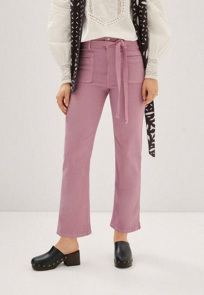 Różowe dżinsy o prostym kroju, z wiązaniem w talii i dwoma przednimi kieszeniami, w połączeniu z czarnymi klapkami. Tekstura jest gładka i miękka.