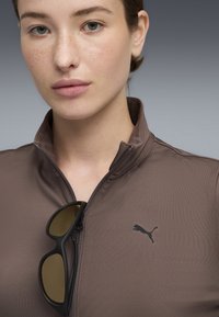 Brun zip-jakke lavet af glat stof, med et sort Puma-logo på venstre side og sorte solbriller hvilende på kraven.