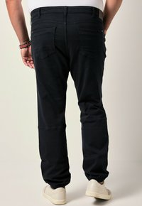 Uomo che indossa pantaloni jogger neri con polsini elasticizzati e scarpe da ginnastica bianche, visto da dietro su uno sfondo semplice.