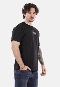 T-shirt nera in cotone con maniche corte, caratterizzata da una piccola grafica bianca sul petto. Abbinata a jeans in denim scuro.