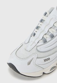 Chaussure de sport blanche avec une tige en mesh, des accents gris et un bout rond. Présente un logo bien en vue et une semelle texturée pour une meilleure adhérence.