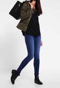 Olivengrøn quilted jakke, sort top, blå skinny jeans og sorte ankelstøvler. Modellen står med en sort tekstureret håndtaske på sin skulder.