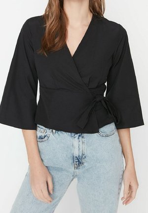 Frau trägt eine schwarze Wickelbluse mit dreiviertellangen Ärmeln, die an der Taille gebunden ist, kombiniert mit hellblauen, hoch taillierten Jeans.