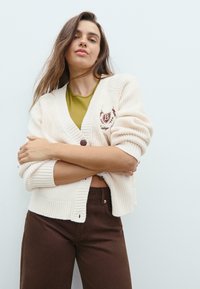 Gilet en crème texturé avec col en V, poignets côtelés, fermeture à boutons et logo brodé sur la poitrine, associé à un haut vert olive et un pantalon marron.