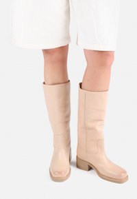 Μπότες από δερμάτινο υλικό σε μπεζ απόχρωση μέχρι το γόνατο, με λεία υφή, ελαφριά γυαλάδα, τετράγωνες μύτες και τακούνι με εσοχή. Συνδυάζονται με λευκές βερμούδες.