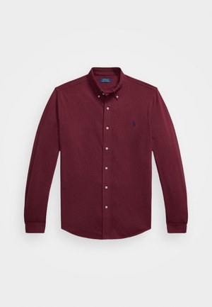 Sarkanais veļa ar garām piedurknēm, ar apkakli, priekšējiem pogām un mazu tumši zilu izšuvumu kreisajā krūšu daļā.