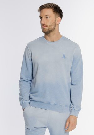 Mężczyzna w jasnoniebieskiej bluzie i pasujących spodniach, stojący z jedną ręką w kieszeni, patrzący w lewo.