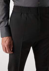 Homme portant un pantalon noir à taille élastique et une chemise grise à manches longues avec poignets boutonnés, debout avec la main gauche près de la poche.