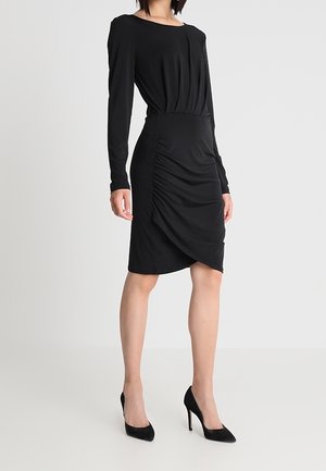 Kobieta w czarnej sukience z marszczeniami na długim rękawie stoi w czarnych wysokich obcasach na jednolitym białym tle.