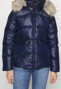 Mörkblå pufferjacka med dragkedja fram, quiltat mönster och fuskpälskant på huvan. Har dragkedjefickor på sidorna och ribbade muddar.