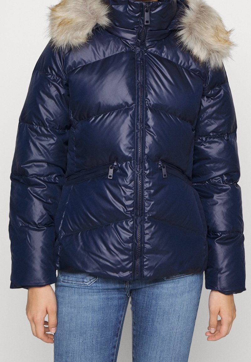Mörkblå pufferjacka med dragkedja fram, quiltat mönster och fuskpälskant på huvan. Har dragkedjefickor på sidorna och ribbade muddar.