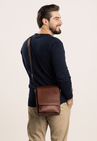 Braune Leder-Umhängetasche mit Klappenverschluss, verstellbarem Schulterriemen, glatter Textur und minimalistischen Design, die über der Schulter getragen wird.