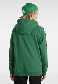 Sudadera verde con un bolsillo tipo canguro, capucha con cordón y rayas gráficas negras en las mangas. Combinada con una gorra verde de ala plana.