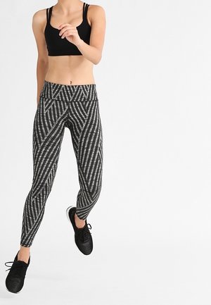 Femme en brassière de sport noire et leggings rayés noir et gris, en pleine foulée comme si elle faisait du jogging ou de l'exercice, portant des chaussures de sport noires.