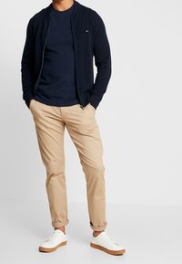 Námornícká bomber bunda na zip přes námořnické tričko, doplněná béžovými chino kalhotami a bílými teniskami s hnědými detaily. Jednoduchý, texturovaný design.