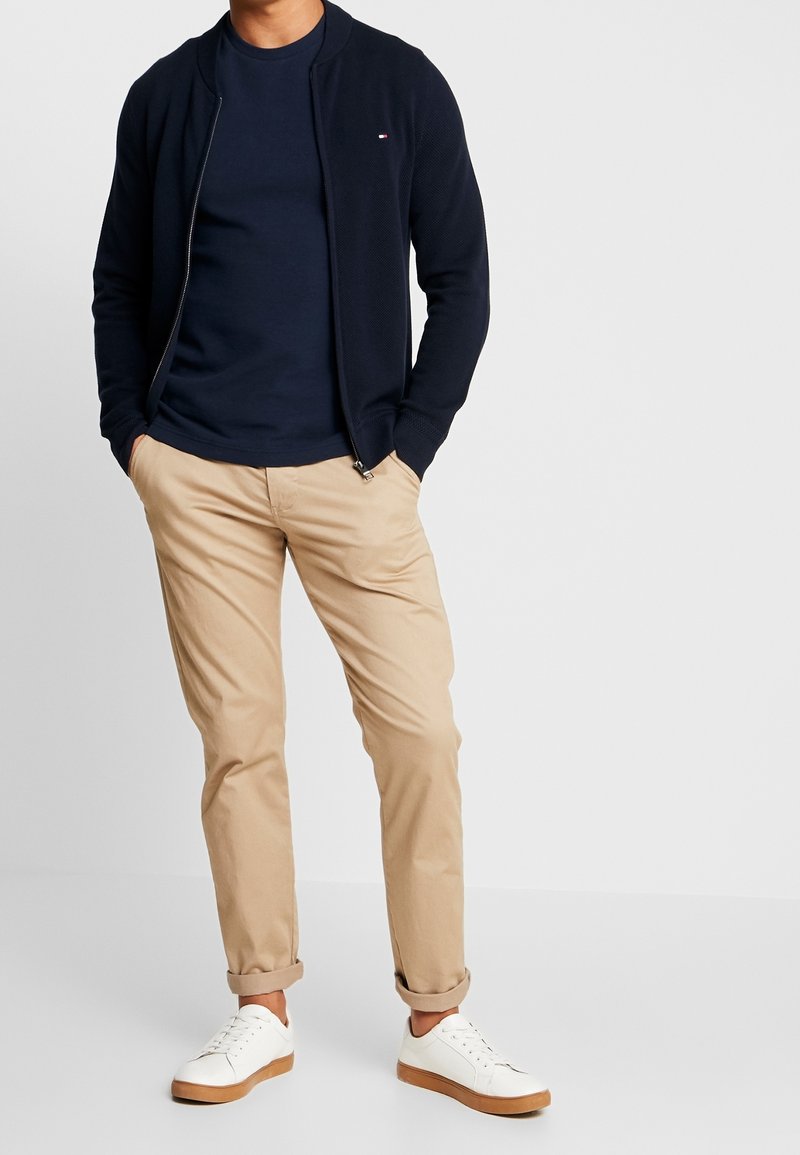 Námornícká bomber bunda na zip přes námořnické tričko, doplněná béžovými chino kalhotami a bílými teniskami s hnědými detaily. Jednoduchý, texturovaný design.