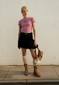 Ružičasta kratka majica s logotipom, tamno traper suknja s naborima, čarape do koljena od čipke, bež čizmice s vezicama, smeđa torbica s privjeskom.