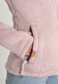 Giacca rosa lavorata a maglia caratterizzata da una texture morbida, maniche lunghe, tasche laterali e un cordoncino con un accento di perlina in legno.