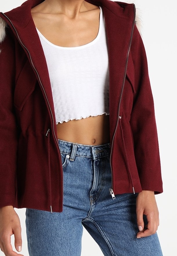 Burgunderfarbene Zip-Jacke mit Kapuze und fellbesetztem Kragen, taillierter Schnitt und zwei Fronttaschen, kombiniert mit einem weißen cropped Top und blauen Jeans.