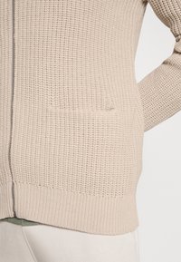 Beżowy sweter robiony na drutach o teksturowej powierzchni, z zamkiem błyskawicznym z przodu i dwiema bocznymi kieszeniami. Dolna krawędź i mankiety są prążkowane.