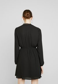 Svart klänning med långa ärmar, insvängd midja och spetsdetaljer vid halsringningen. Lättviktig, flödande tyg, ovan knä-längd.