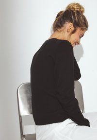 Frau mit blonden Haaren im Dutt, die einen schwarzen Pullover und weiße Hosen trägt, seitlich auf einem Metallstuhl vor einer schlichten Wand sitzt.