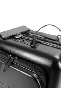 Schwarzer Hartschalenkoffer mit glatter Oberfläche, ausgestattet mit einem eingebauten USB-Anschluss, einem Schloss und einem eleganten Design. Ein Smartphone liegt darauf.